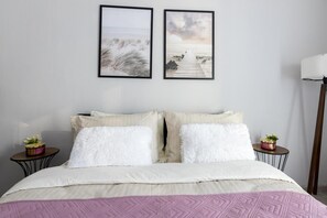 Quarto com cama, duas almofadas, mesa de cabeceira com planta e duas imagens emolduradas na parede.
