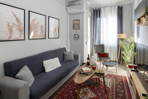 A modern living room with a dark blue sofa, a coffee table, and a red patterned rug. There are framed pictures on the wall, a small side table with a bottle and glasses, and a chair with a red cushion. - Apartment 'George House' with Balcony, Wi-Fi and Air Conditioning (Θεσσαλονίκη)