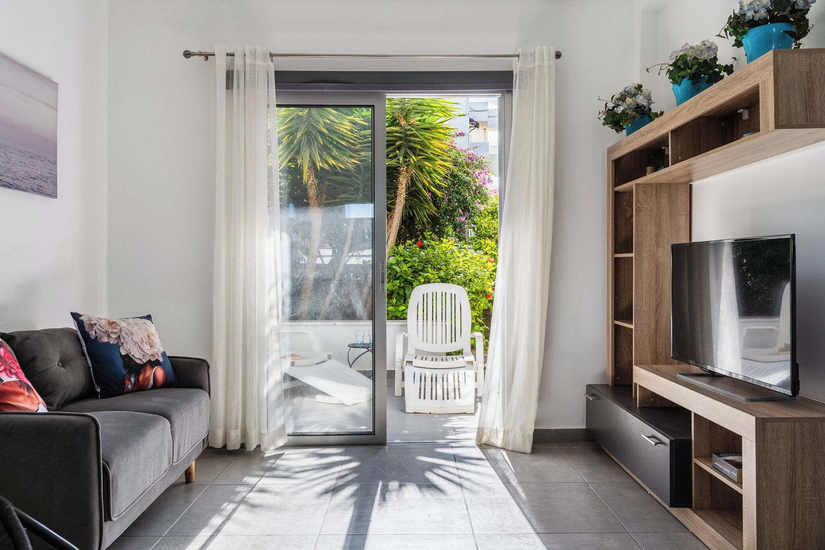 A modern living room with a sofa, a TV on a wooden stand, a sliding glass door leading to a balcony with a chair and plants.