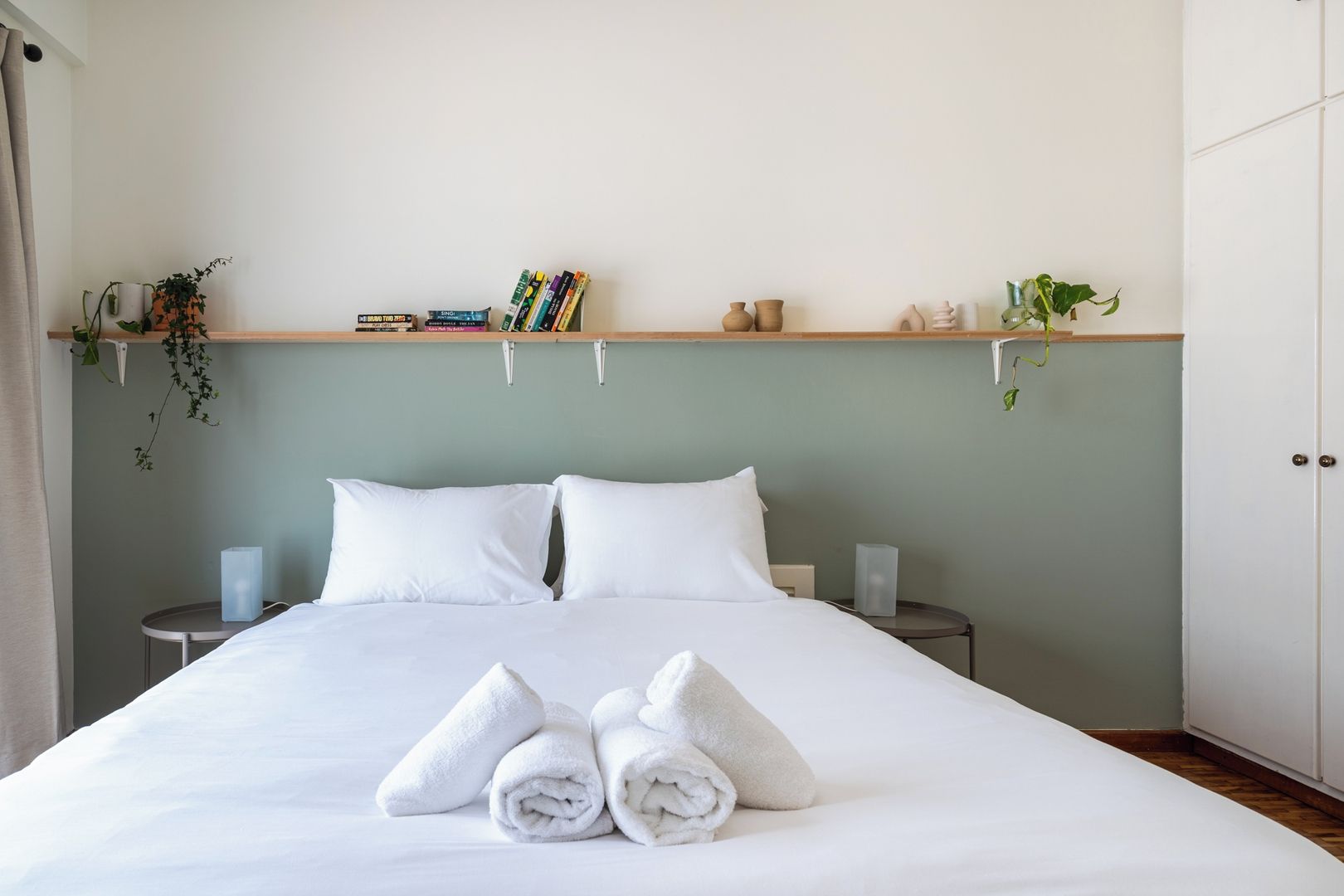 Un letto rifatto con lenzuola e asciugamani bianchi, due comodini con lampade, una mensola con libri e oggetti decorativi, e un armadio.