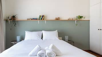 Un letto rifatto con lenzuola e asciugamani bianchi, due comodini con lampade, una mensola con libri e oggetti decorativi, e un armadio.
