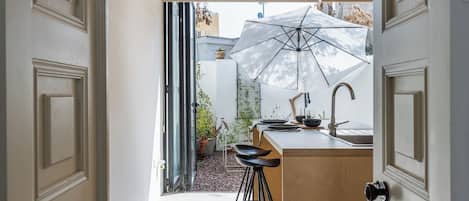 Corredor estreito que dá para uma área de cozinha com um banco, pia e um guarda-sol grande que oferece sombra.