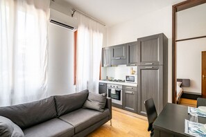 A modern living room with a sofa and a dining table, a kitchenette with grey cabinets, and a window with curtains. - GuestHost - Padova Suite Isabella 500m from the Duomo! (Padova)