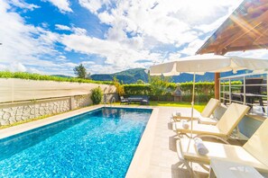 Una piscina con sillas de descanso y sombrillas, rodeada por un muro de piedra y vegetación, con montañas al fondo.