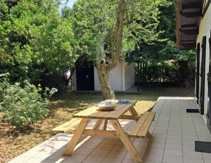 A wooden picnic table with benches, a bowl, and a small table in a garden area with trees and a building in the background. - Family home 500 meters from the beach, sleeps 6 (Andernos-les-Bains)