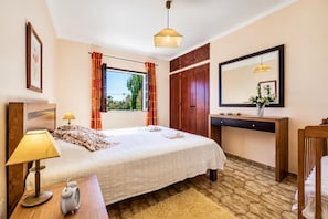 Ein Hotelzimmer mit Bett, Nachttisch, Lampe, Wecker, Fenster mit Vorhängen, Holzschrank, Spiegel und einem kleinen Schreibtisch mit einer Vase.