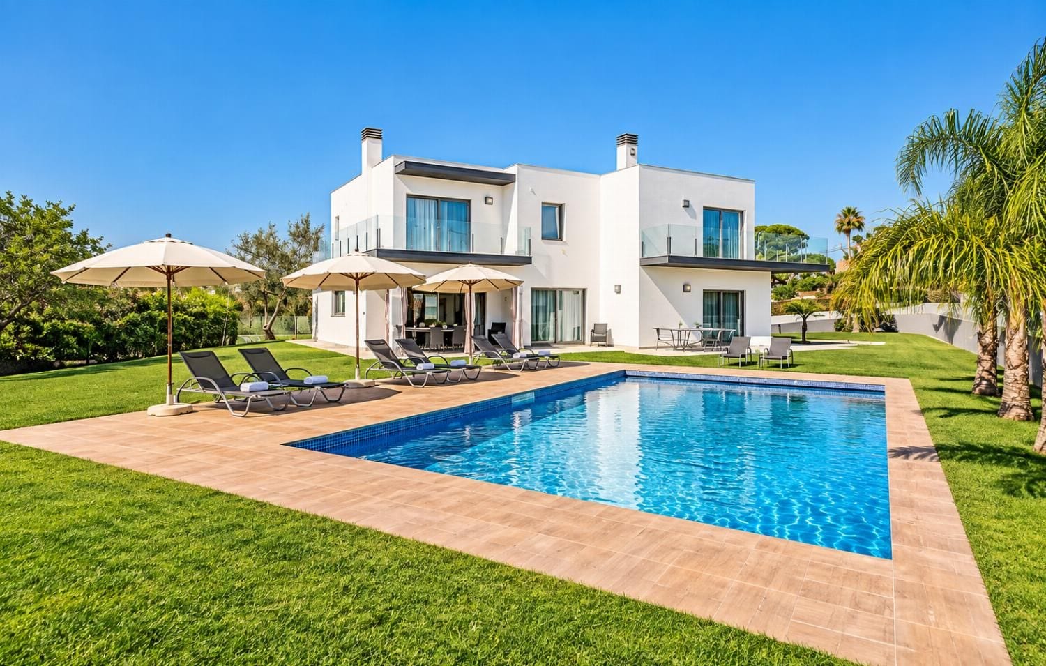 A modern two-story house with a swimming pool, sun loungers, and outdoor umbrellas.
