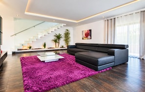 A modern living room with a black leather sofa, a glass coffee table, and a vibrant purple rug. A staircase with glass railings is visible in the background. - Villa Dala - Four Bedroom Villa, Sleeps 8 (Albufeira)