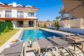 Zona de piscina amb gandules, taula amb ampolla i gots, i edifici amb façana de pedra i teulada vermella.