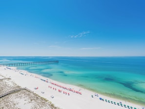 A beach with a pier extending into the water, clear turquoise water, and a row of beach umbrellas. - The Pearl of Navarre #503 | Gulf Front Condo + Beach Views (Navarre Beach)