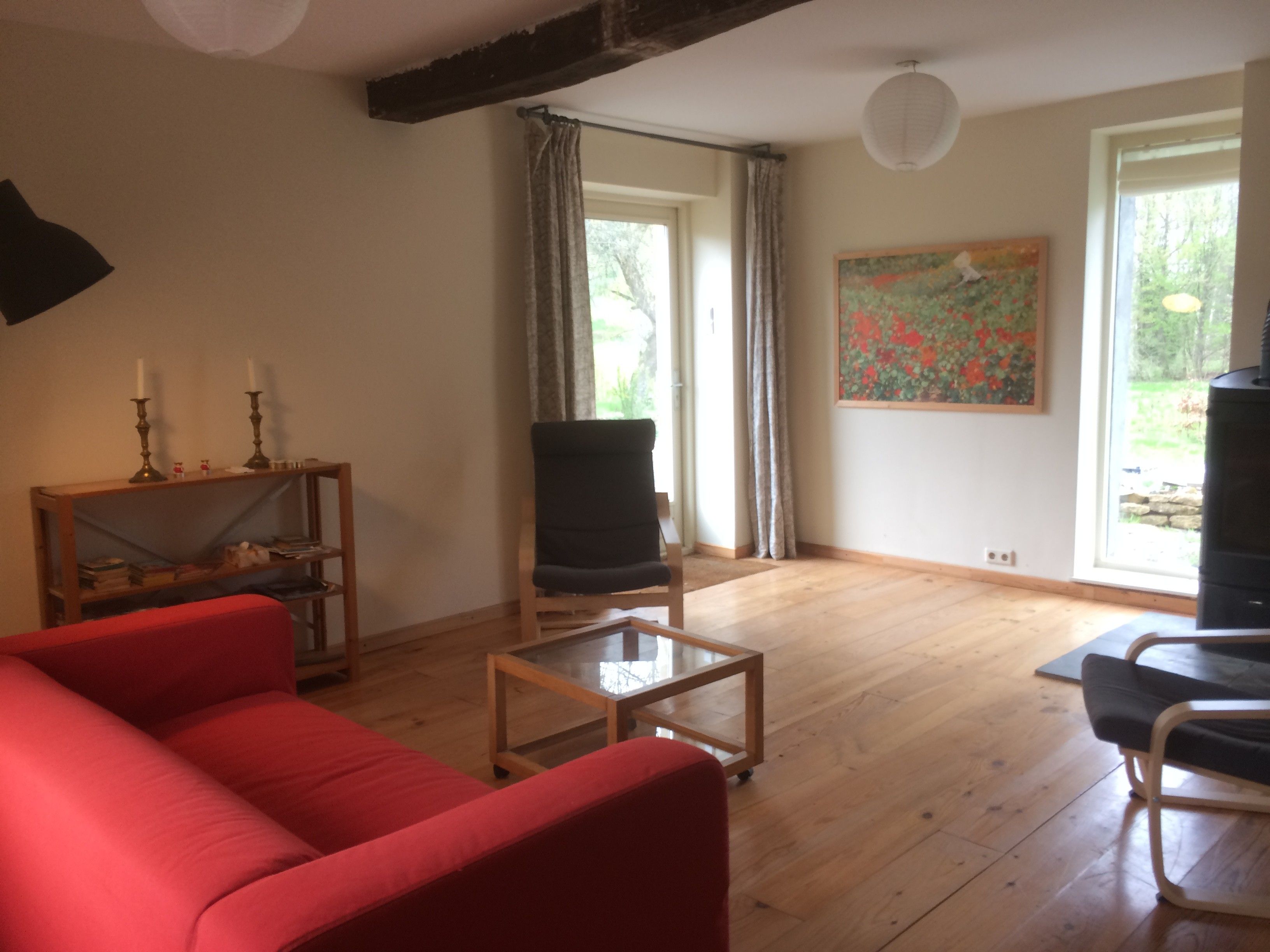 A living room with a red sofa, a black armchair, a glass coffee table, and a wooden bookshelf. There is a painting on the wall and a window with curtains.
