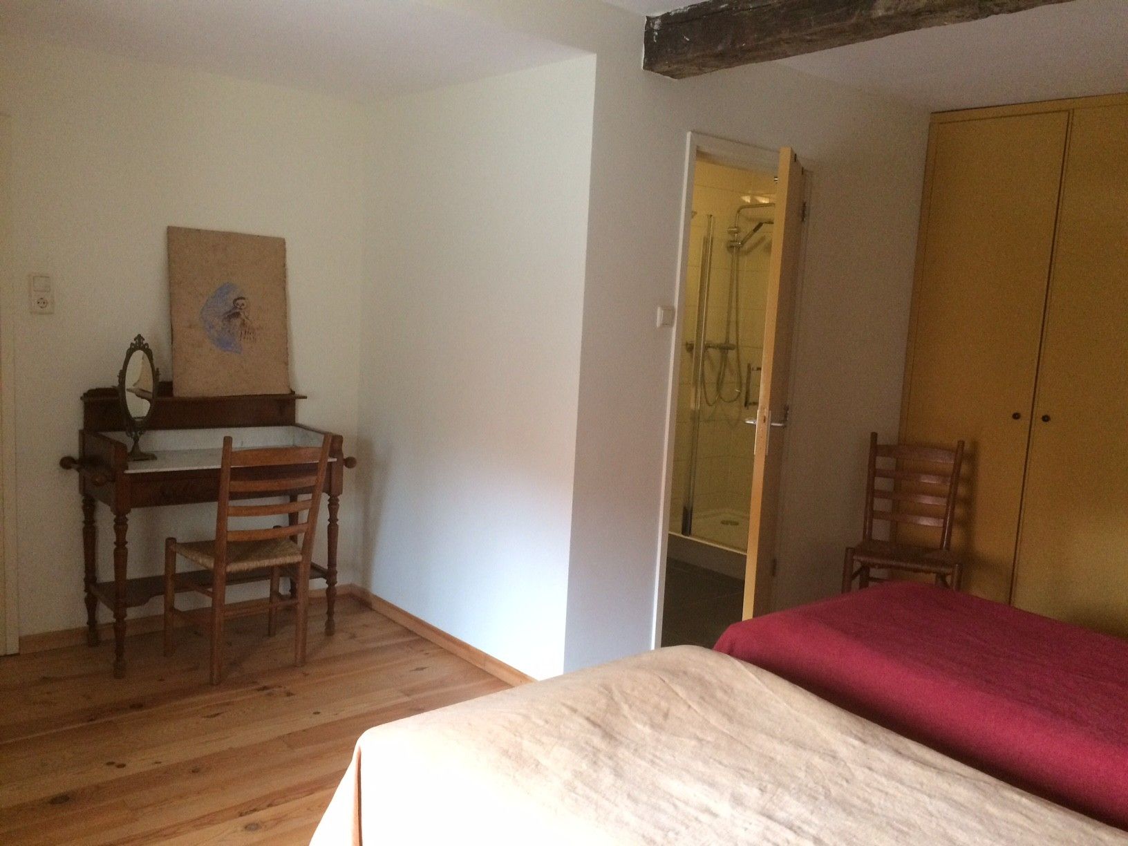 Een kamer met een houten bureau, twee stoelen, een bed met een rode spreidek en een kledingkast.