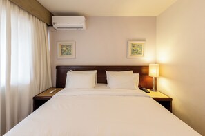 Ein Hotelzimmer mit einem großen Bett, zwei Nachttischen, einem Beistelltisch mit Lampe, einer Klimaanlage und zwei gerahmten Kunstwerken an der Wand.