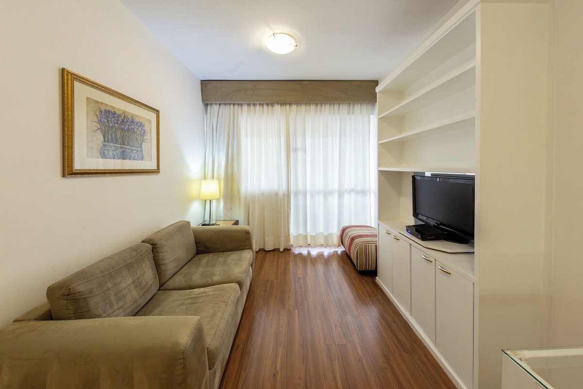 Ein Wohnzimmer mit einer Couch, einem Fernseher auf einem Schrank, einer Lampe und einem Fenster mit Vorhängen.