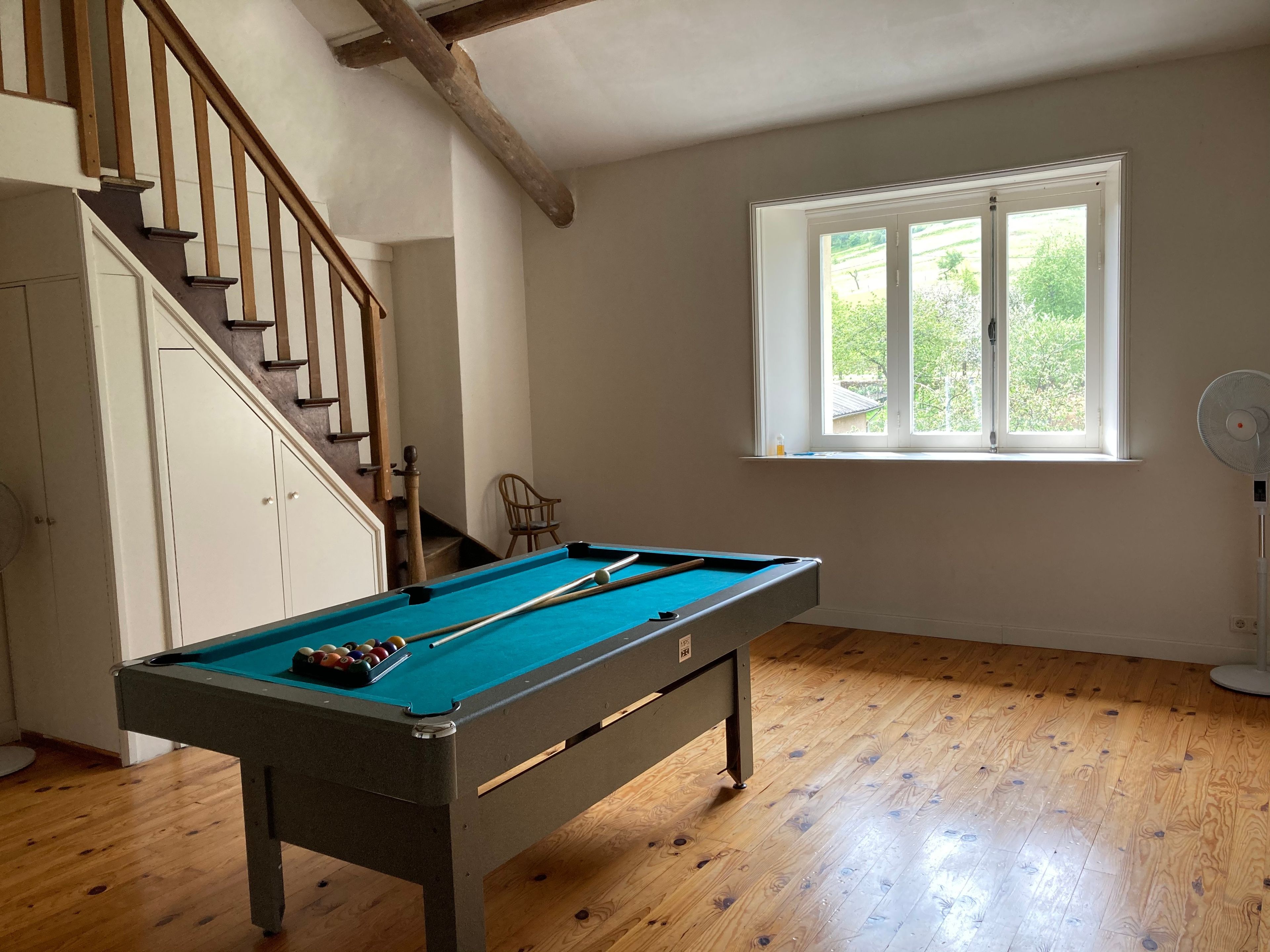 A room with a pool table, wooden flooring, a window with a view, and a staircase.