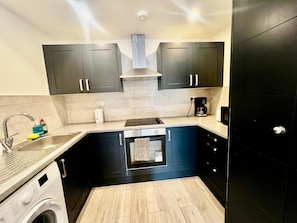 A compact kitchen with dark cabinets, a built-in oven, a coffee maker, and a washing machine. - Apartment Sleeps 5 Near High Street (Croydon)