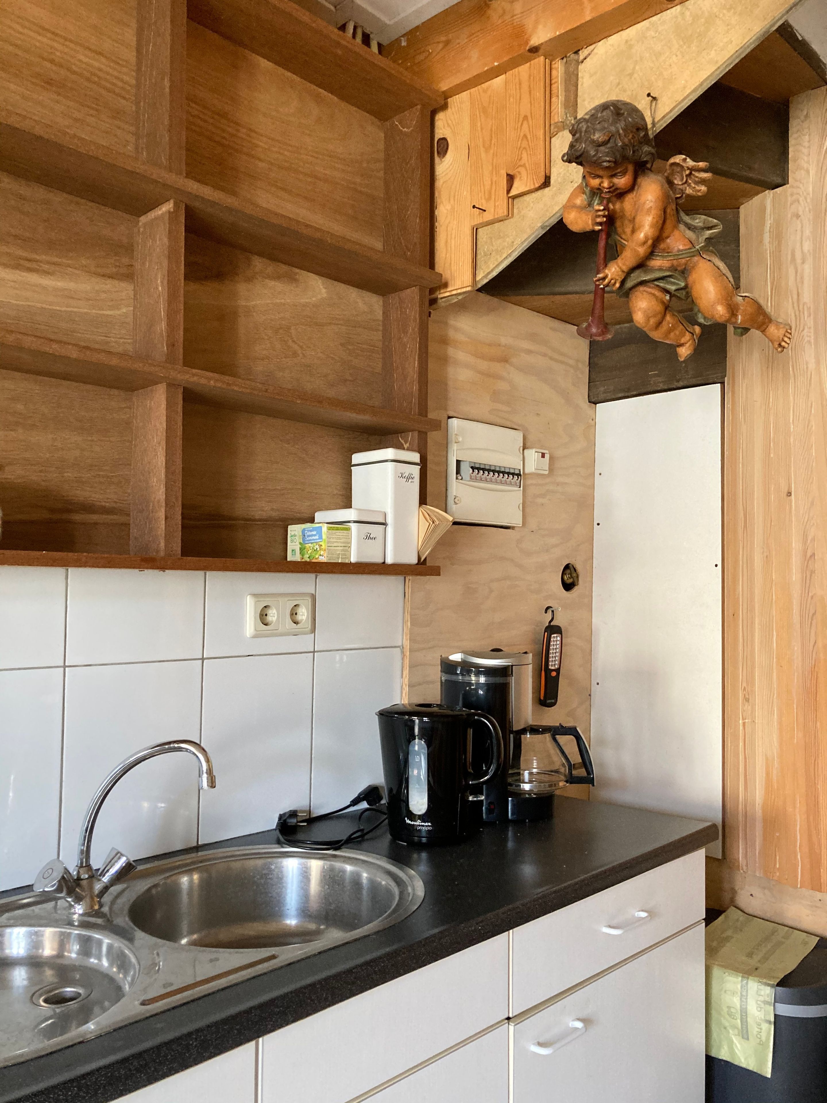 Una cocina con un lavamanos de acero inoxidable, una cafetera y un hervidor. Hay un estante de madera con una figurita decorativa de ángel.