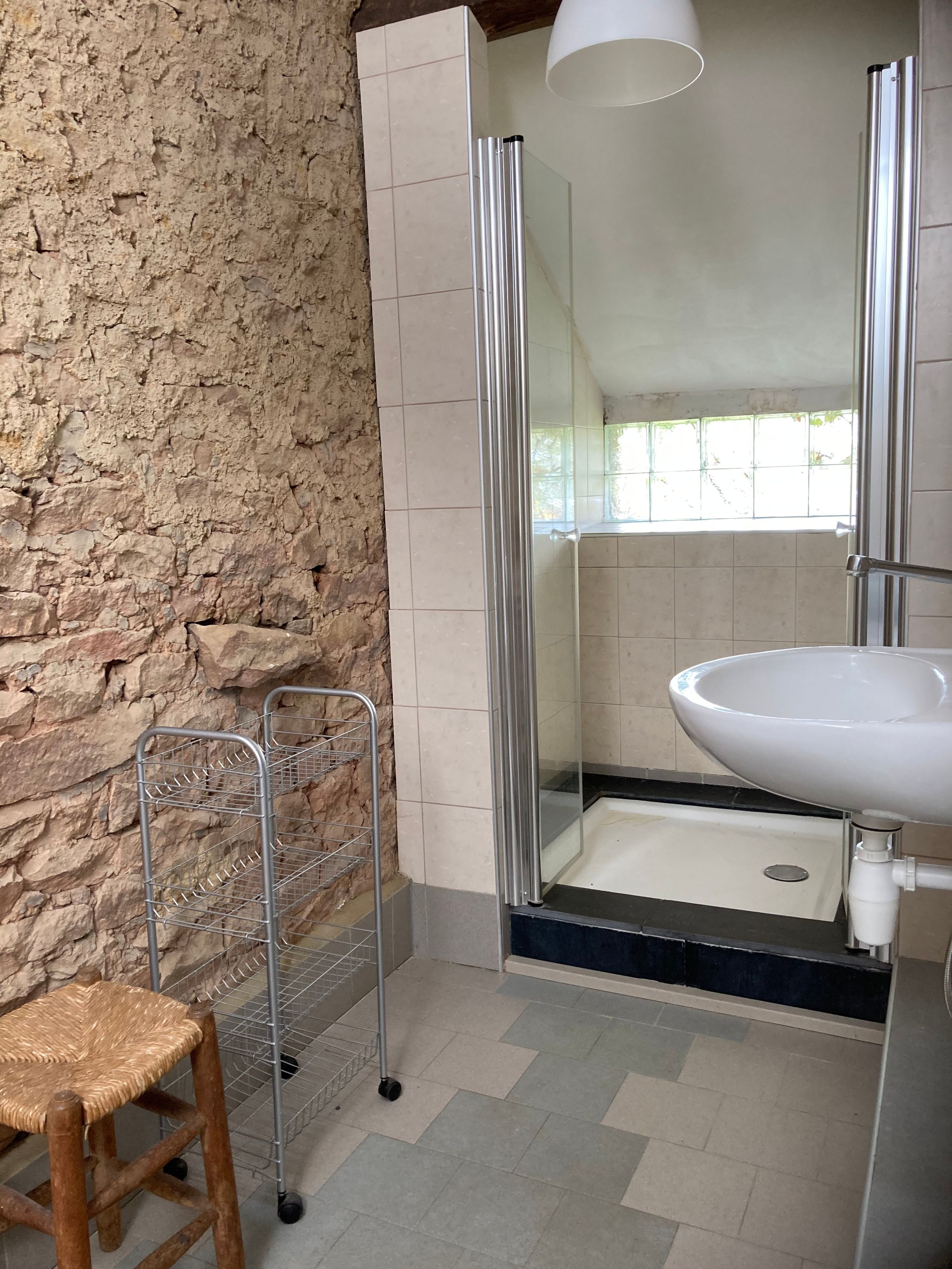 Une salle de bain avec un mur en pierre, une douche, un lavabo et un tabouret.