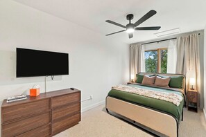 Ein Schlafzimmer mit einem Bett, einem an der Wand befestigten Fernseher, einem Deckenventilator und einem Fenster mit Vorhängen.
