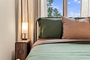 A bed with a green bedspread and two pillows, a wooden nightstand with a lamp, and a window with curtains. - The Cardinal | Wooded Stay Near Downtown + Trails (Bentonville)