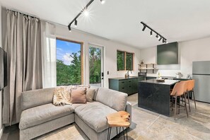 A modern living room with a sectional sofa, a wooden coffee table, and a view of greenery through the windows. - The Cardinal | Wooded Cabin Stay Near Downtown + Trails (Bentonville)