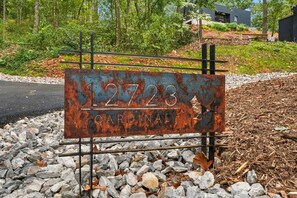 Ein verrostetes Metallschild mit der Nummer '12723' und dem Wort 'CARDINAL' darauf, auf einer Schotterfläche vor Bäumen und einem Gebäude im Hintergrund platziert.