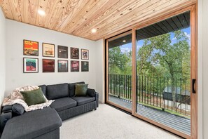 Un soggiorno moderno con un divano modulare grigio, un soffitto in legno e un'ampia porta scorrevole in vetro che dà su un balcone con vista sugli alberi.
