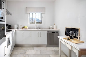 A modern kitchen with white cabinetry, a stainless steel dishwasher, a window with a view, and a chalkboard with glasses and a bottle. - Casa Alborán | Jacuzzi | Billiard | BBQ | Sea view (Málaga)