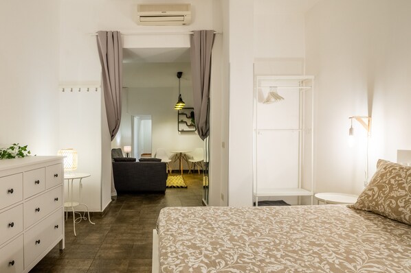 Ein Schlafzimmer mit einem Bett, einem Schminktisch, einem Nachttisch, einer Lampe, einem Spiegel und einer Tür zu einem weiteren Raum.