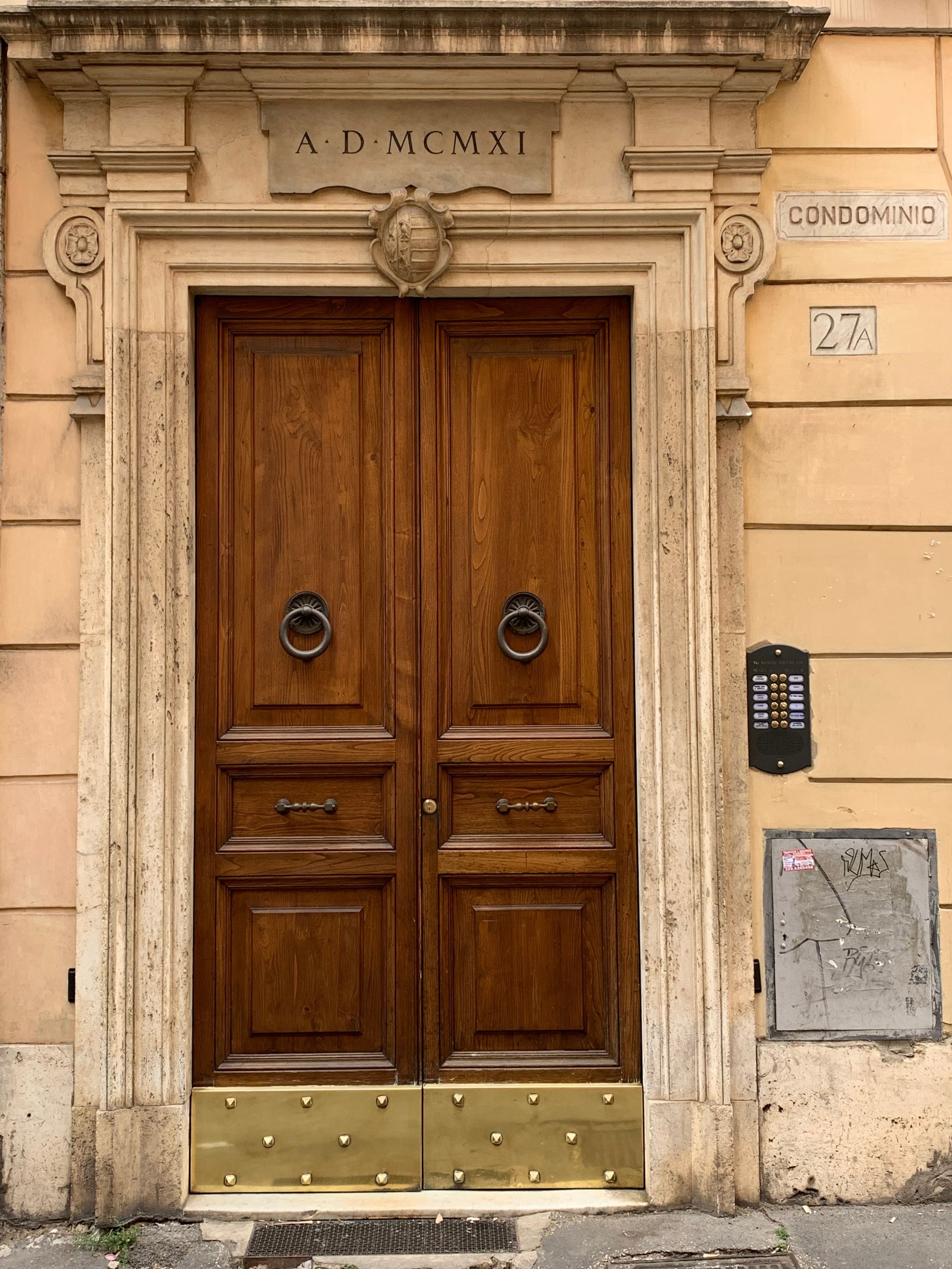 A double wooden door with a brass plate above it, a metal plaque to the right, and a number '27A' on the wall.