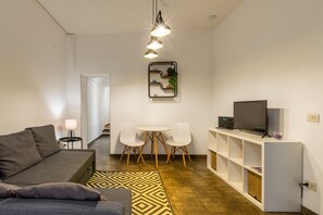 Ein modernes Wohnzimmer mit einer Couch, einem Fernseher auf einem Schrank, einem Essbereich mit Stühlen und einem gemusterten Teppich.