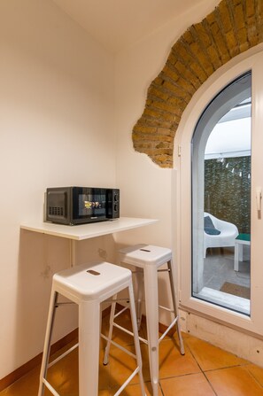 Un coin de pièce avec un mur blanc, une arche en briques, une table blanche sur laquelle se trouve un four à micro-ondes, et deux tabourets blancs.