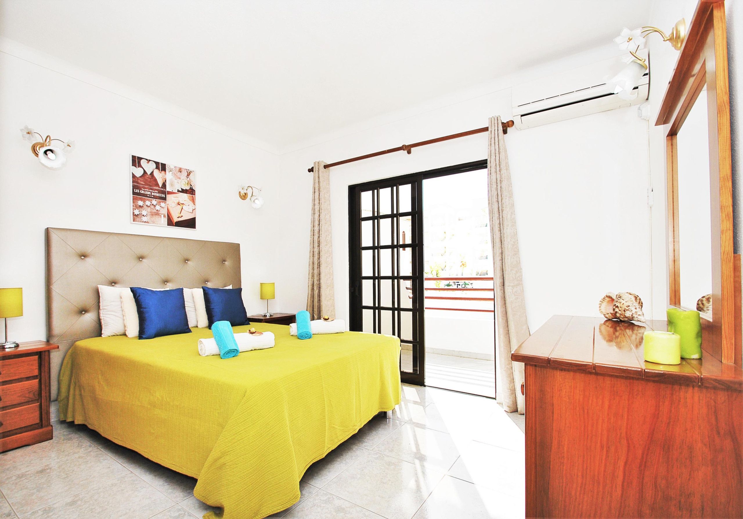 A hotel room with a bed, a nightstand, a dresser, a mirror, and a view of the outside.