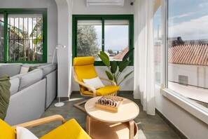 Un salon moderne avec un canapé, un fauteuil jaune, une table ronde sur laquelle est posé un jeu d’échecs, et une grande fenêtre donnant sur des immeubles.