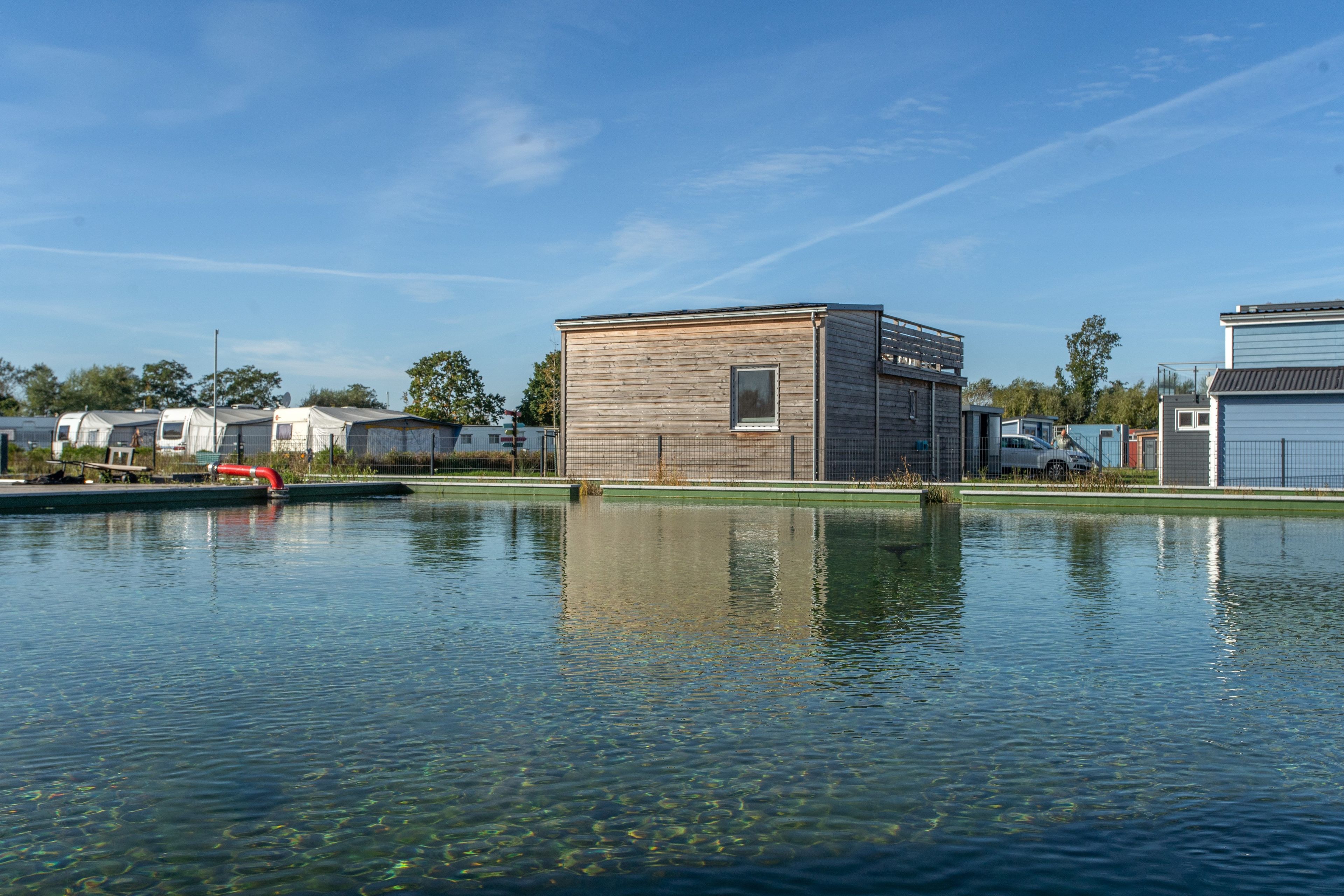 Een woongebied met een helder waterlichaam dat de gebouwen weerspiegelt, een houten constructie en een omheind gebied met stacaravans.