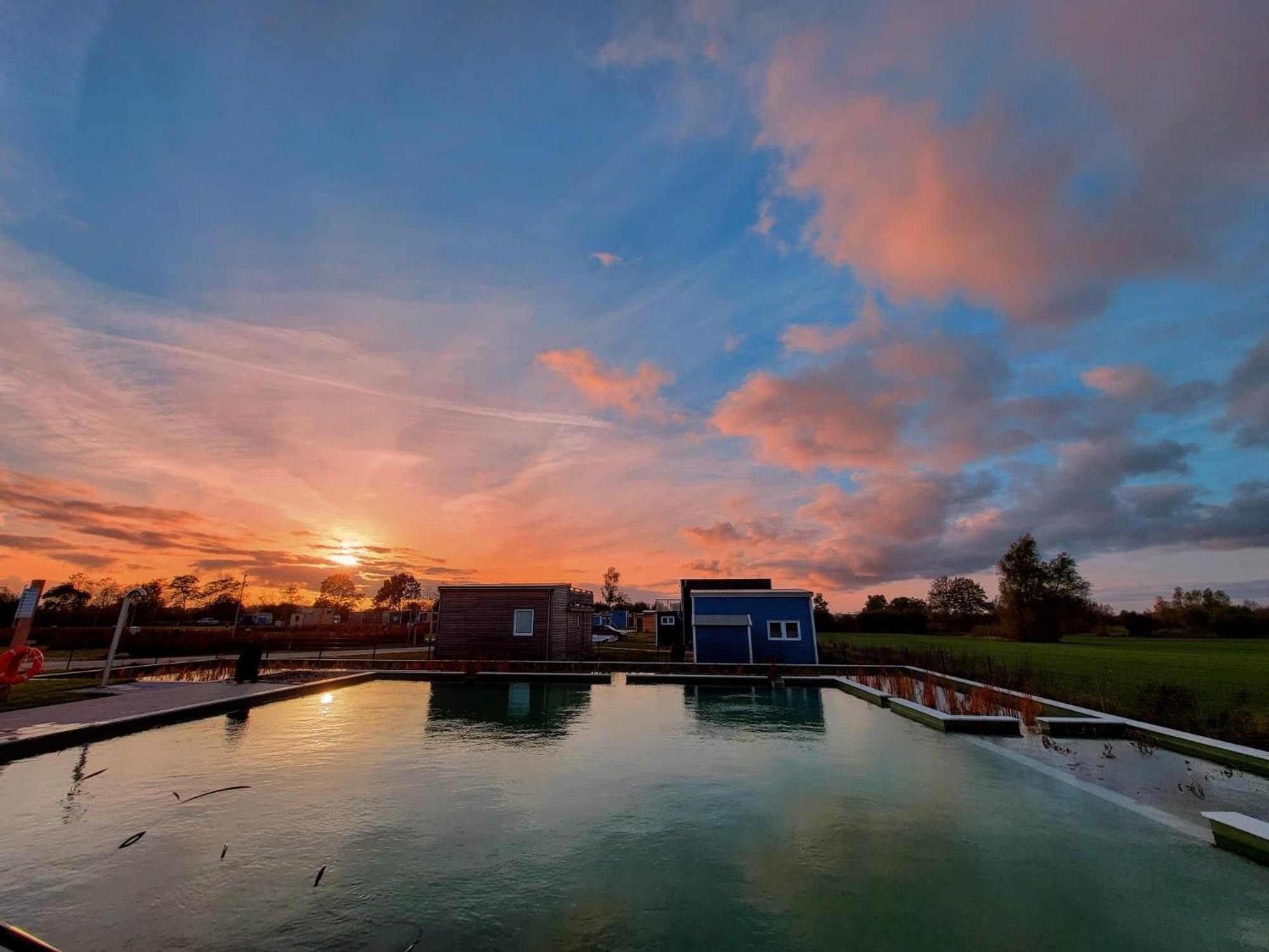 A calm body of water reflecting the sky, surrounded by small buildings and a grassy field.