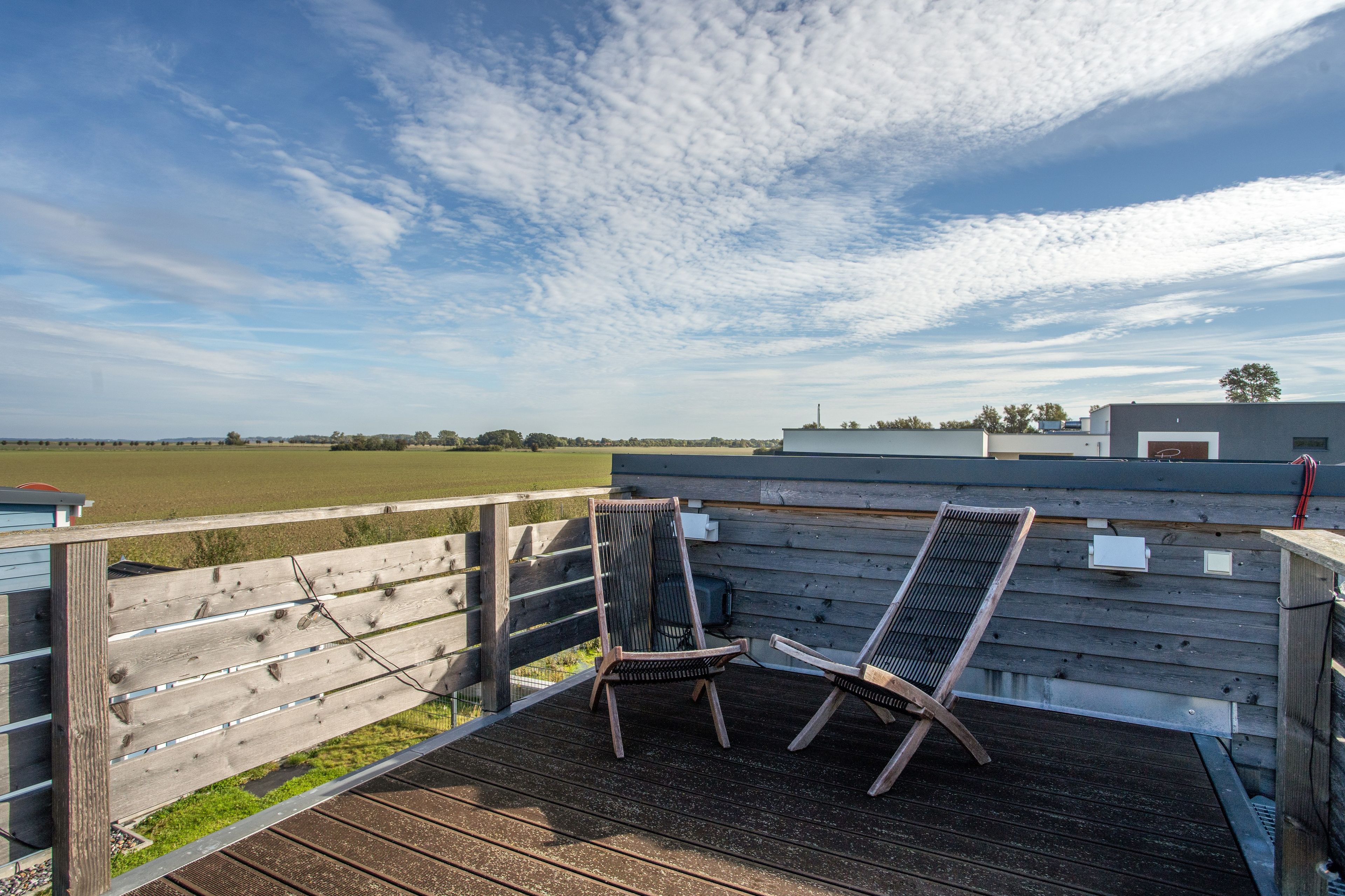Una terraza de madera con dos sillas y una mesa, con vistas a un campo y un edificio.