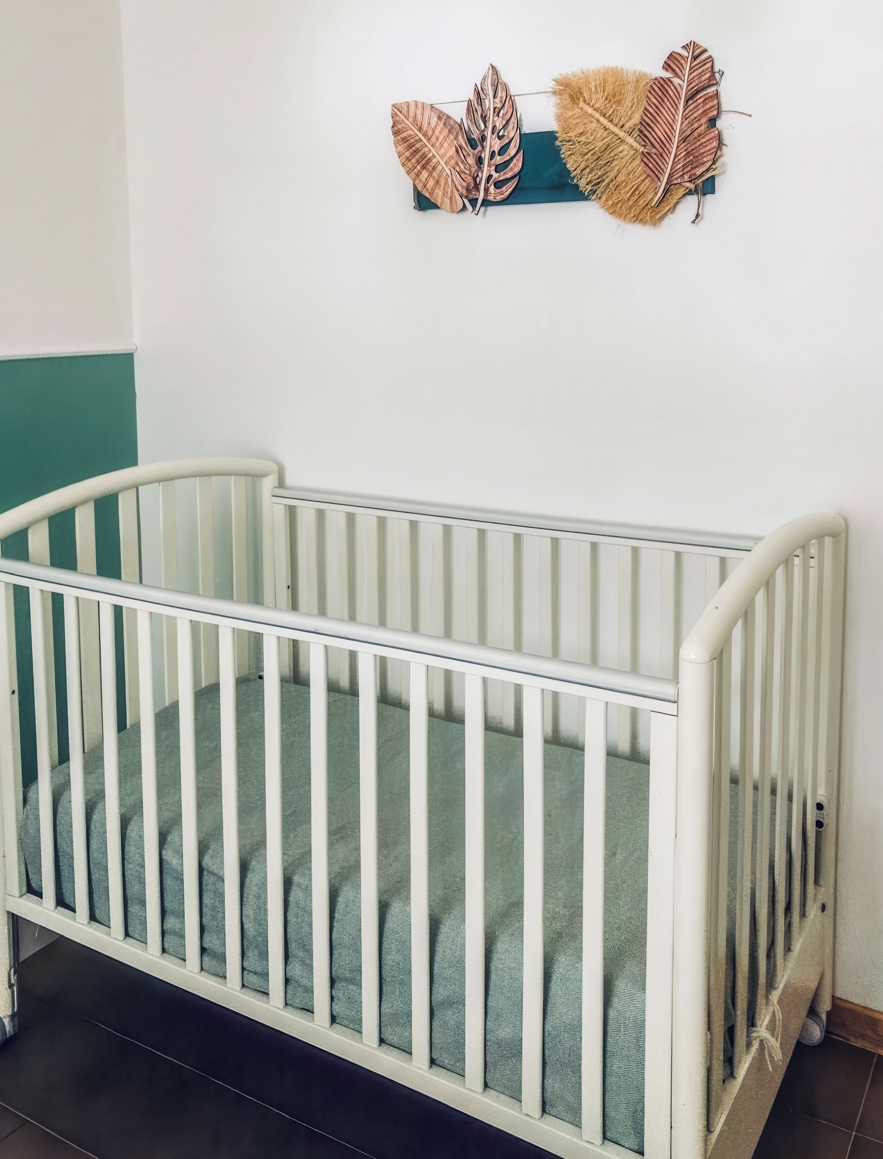 Un berceau blanc avec un matelas vert, placé contre un mur blanc agrémenté d’une touche de couleur turquoise.