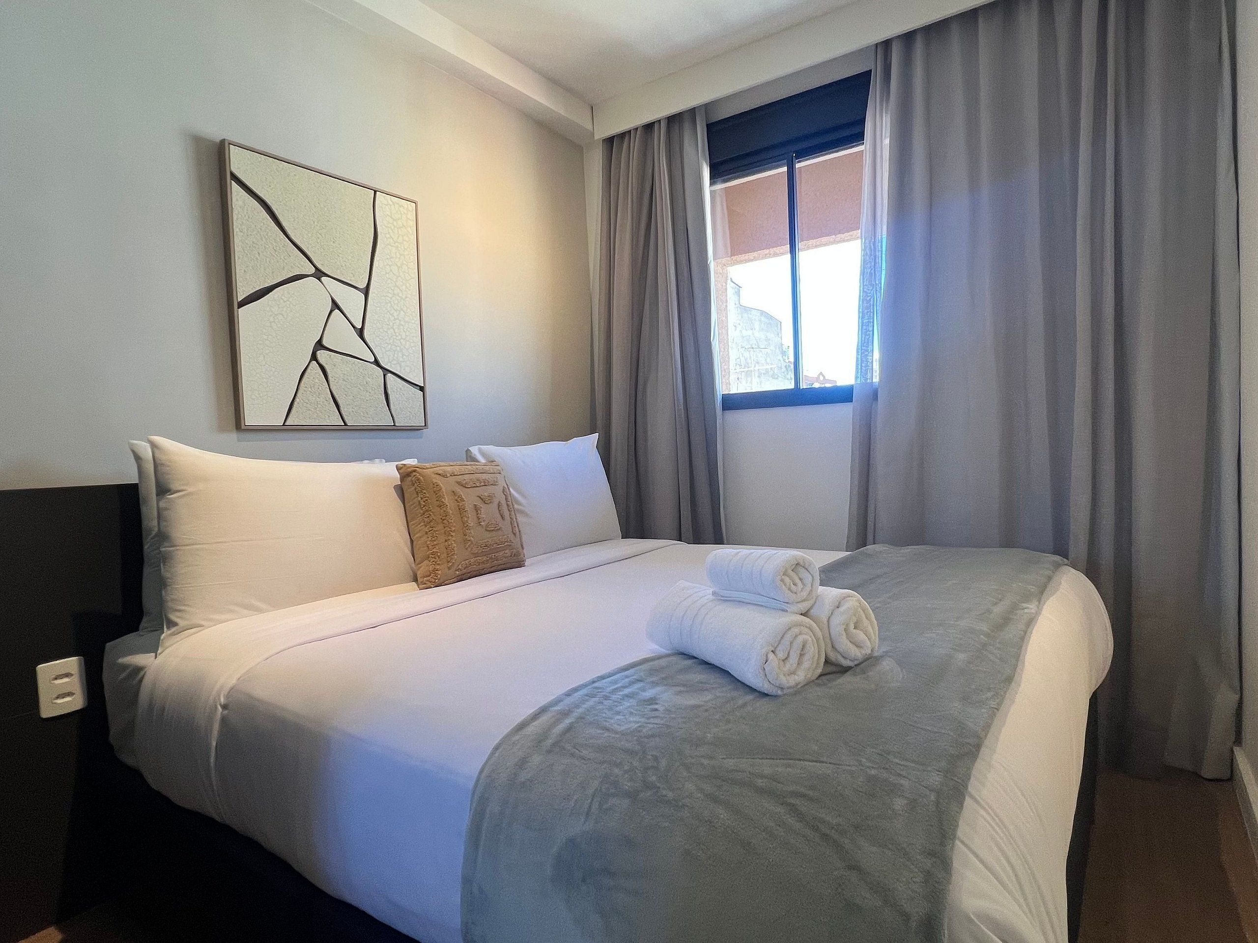 A hotel room with a bed, a pillow, a rolled towel, a window with curtains, and a wall art piece.