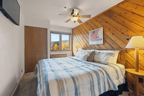 A bedroom with a bed, a window, a ceiling fan, a nightstand with a lamp, and a wooden wall panel. - NEW LISTING: Top Floor 2 Bed 2 Bath Condo. (Breckenridge)