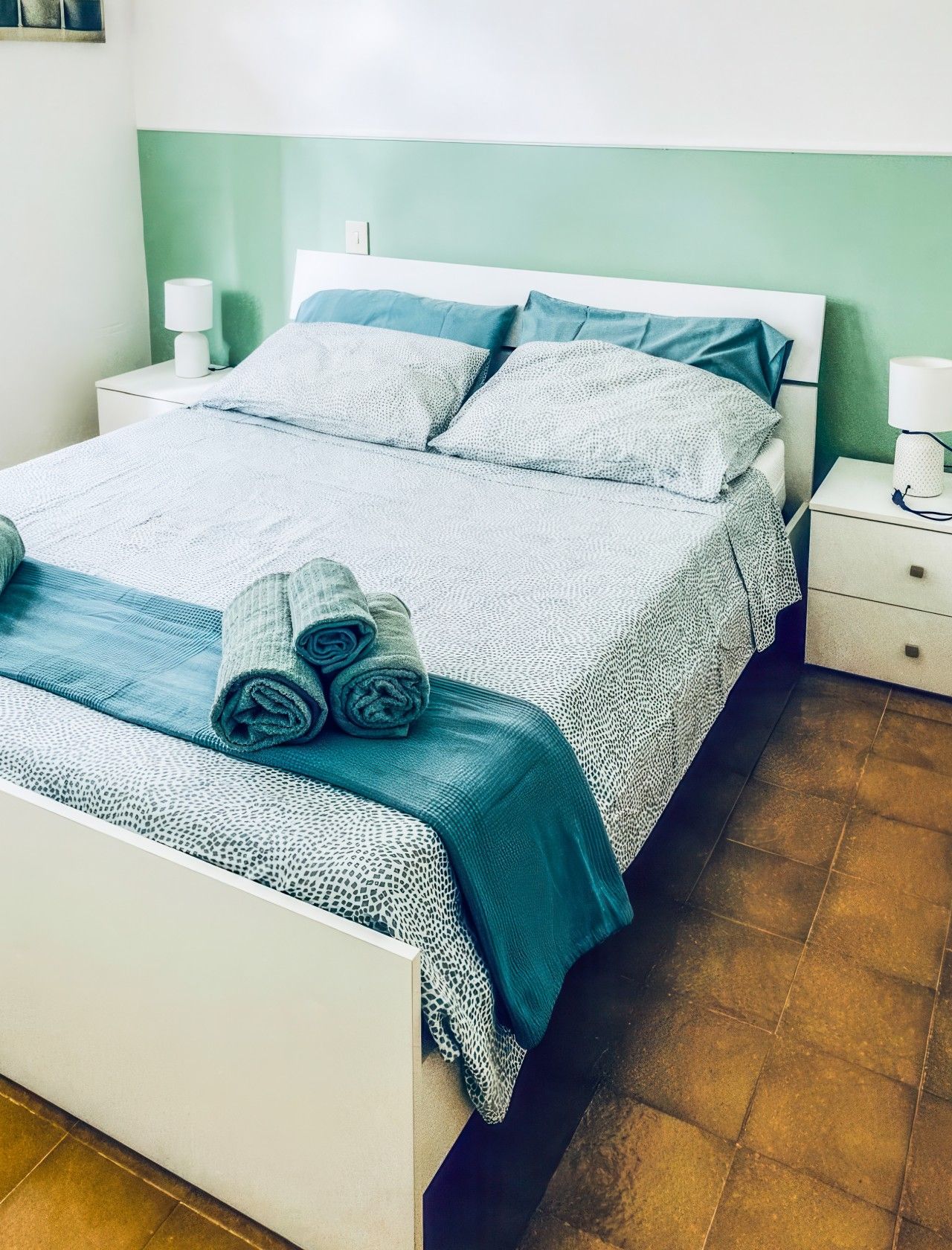 Un lit bien fait, avec des serviettes pliées et une couvre-lit à motifs, une table de chevet avec une lampe et une petite table de nuit également équipée d’une lampe.