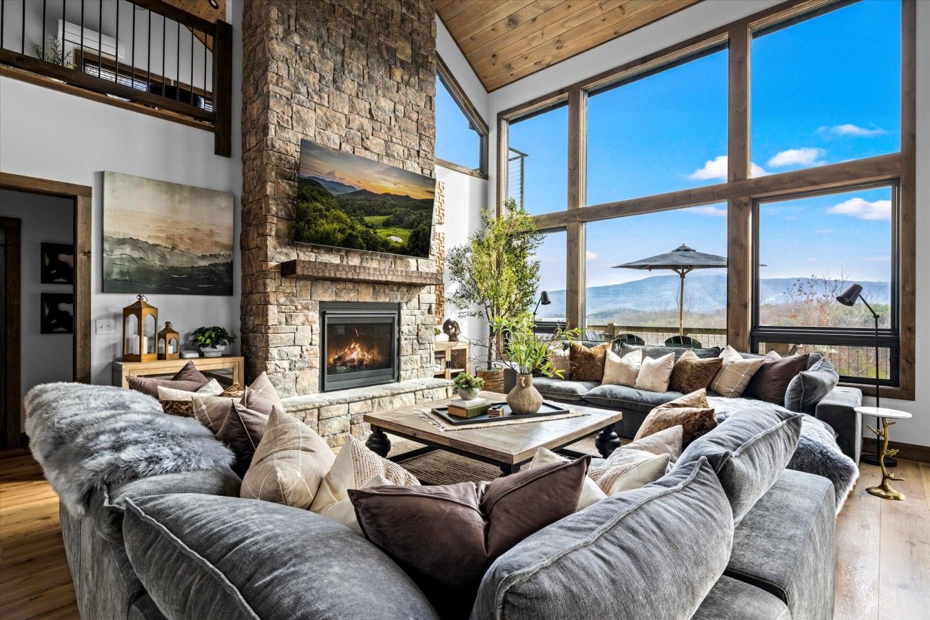 A spacious living room with a stone fireplace, large windows, and a view of the outdoors.