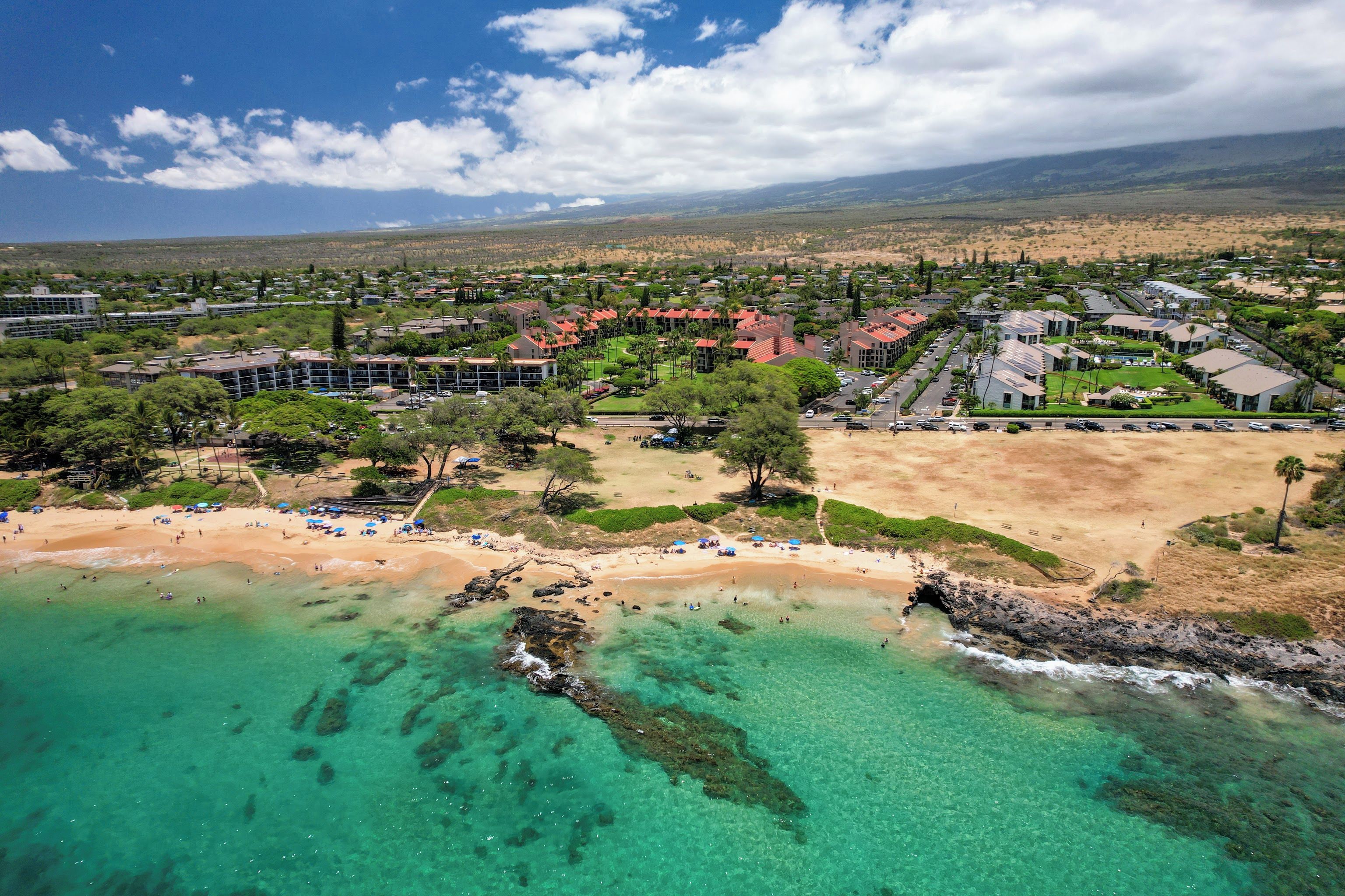 An aerial view of a coastal resort with a beach, clear turquoise waters, and a mountainous backdrop.