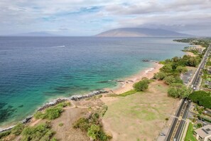 An aerial view of a coastal area with a beach, a road, and a mountain in the distance. - Maui Kamaole E103 by Gather - Garden Condo w/ Two Private Lanais (Kihei)