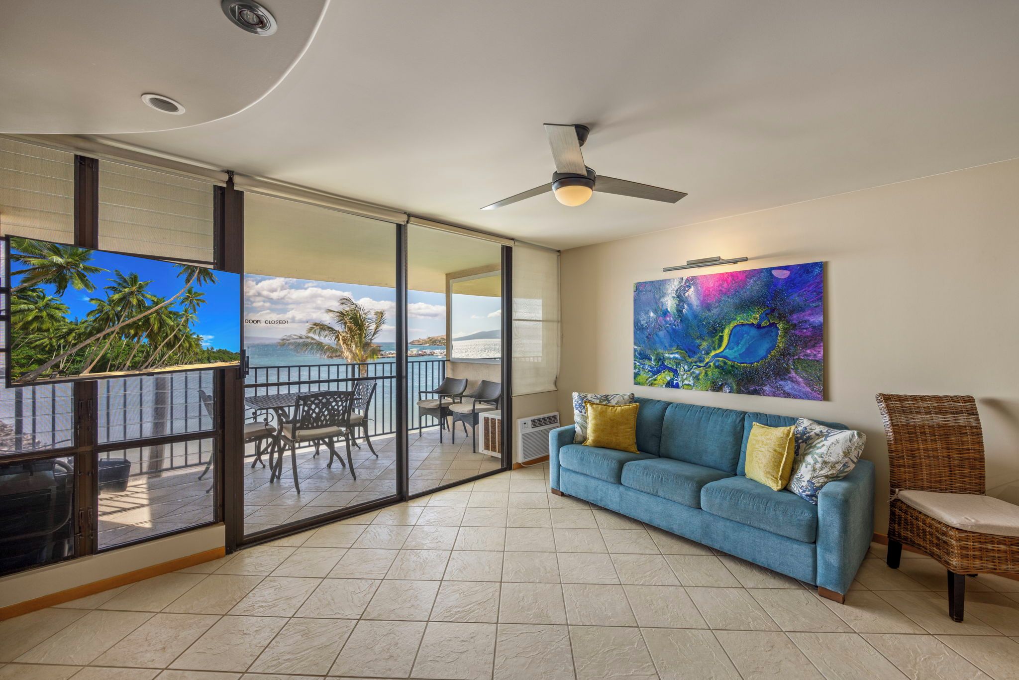 A room with a sliding glass door leading to a balcony, a blue sofa, a ceiling fan, and a large abstract painting.