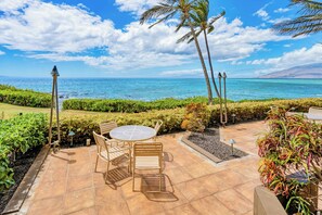 Tiki torches flank the patio table, palms swaying against a wide stretch of blue. - Punahoa Beach 205 by Gather - Oceanfront Condo w/ Private Lanai & Ocean Views (Kihei)