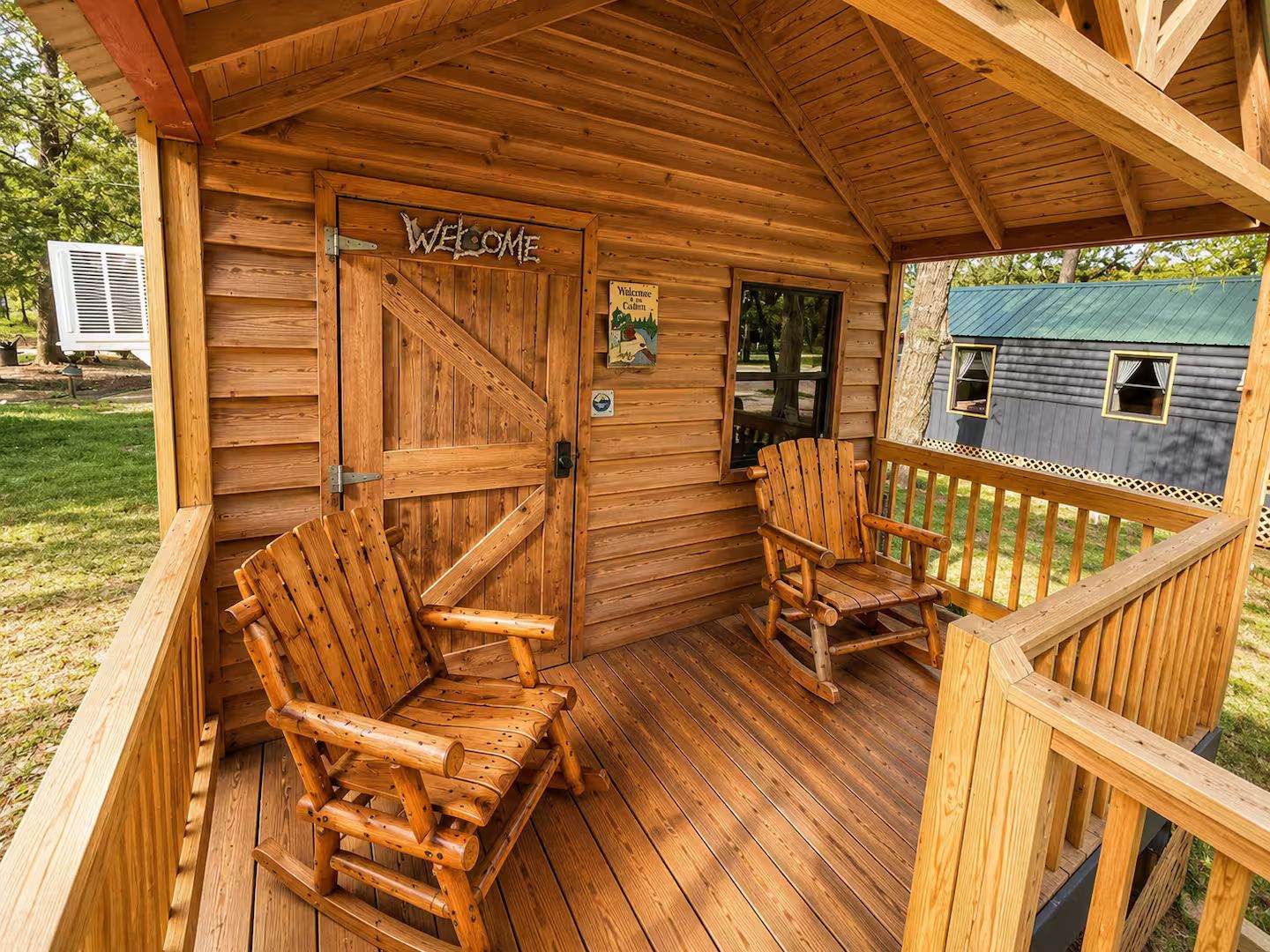 A wooden cabin with a porch, two wooden chairs, a welcome sign, and a small window.