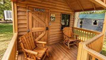 A wooden cabin with a porch, two wooden chairs, a welcome sign, and a small window.