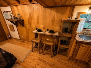 Interior de madera con una mesa de comedor preparada para dos, una zona de cocina con un fregadero y un extintor en el suelo.
