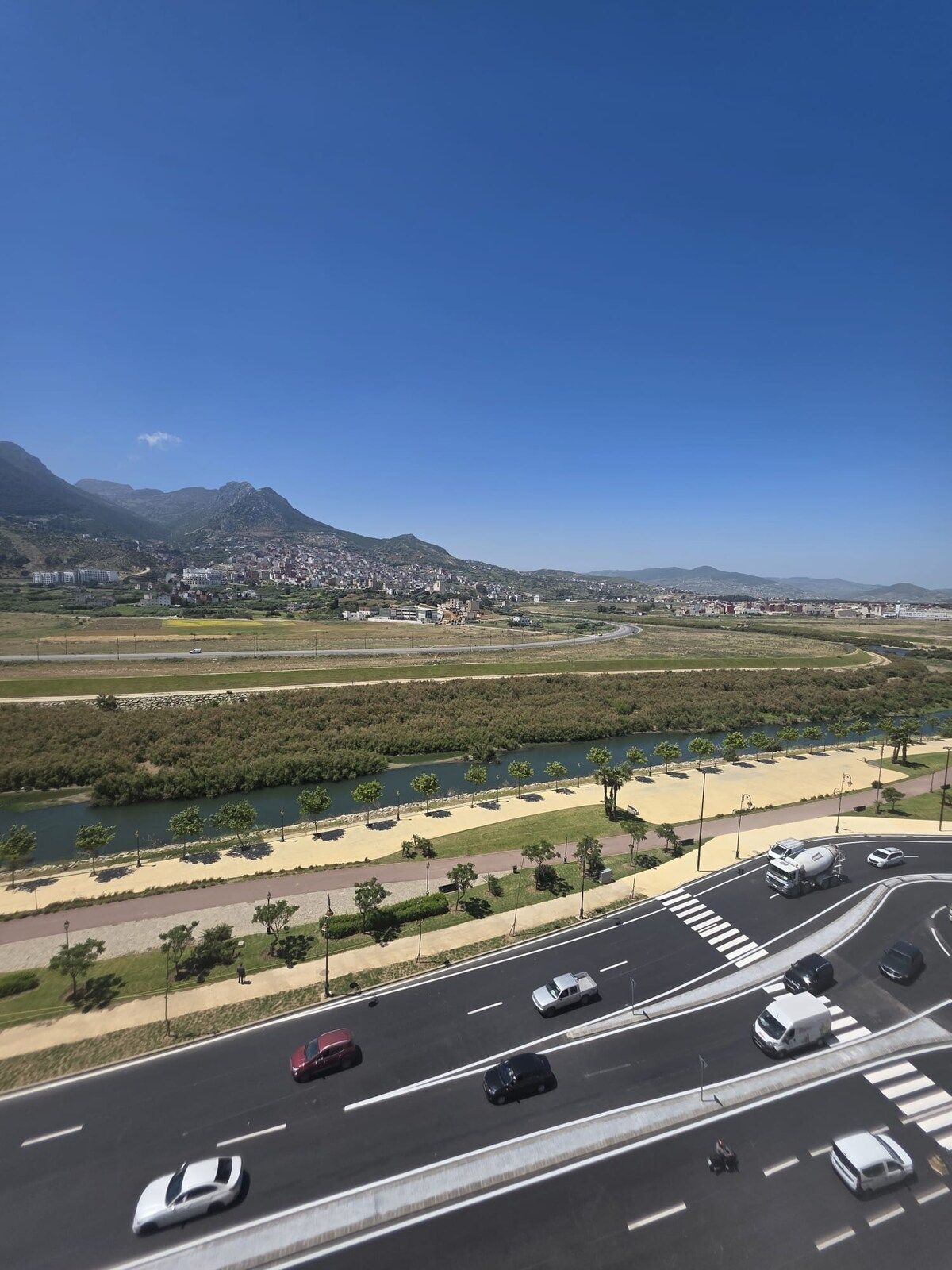 Uma autoestrada com várias faixas e veículos, um rio com árvores e uma passarela para pedestres, e uma paisagem urbana distante com edifícios e montanhas.